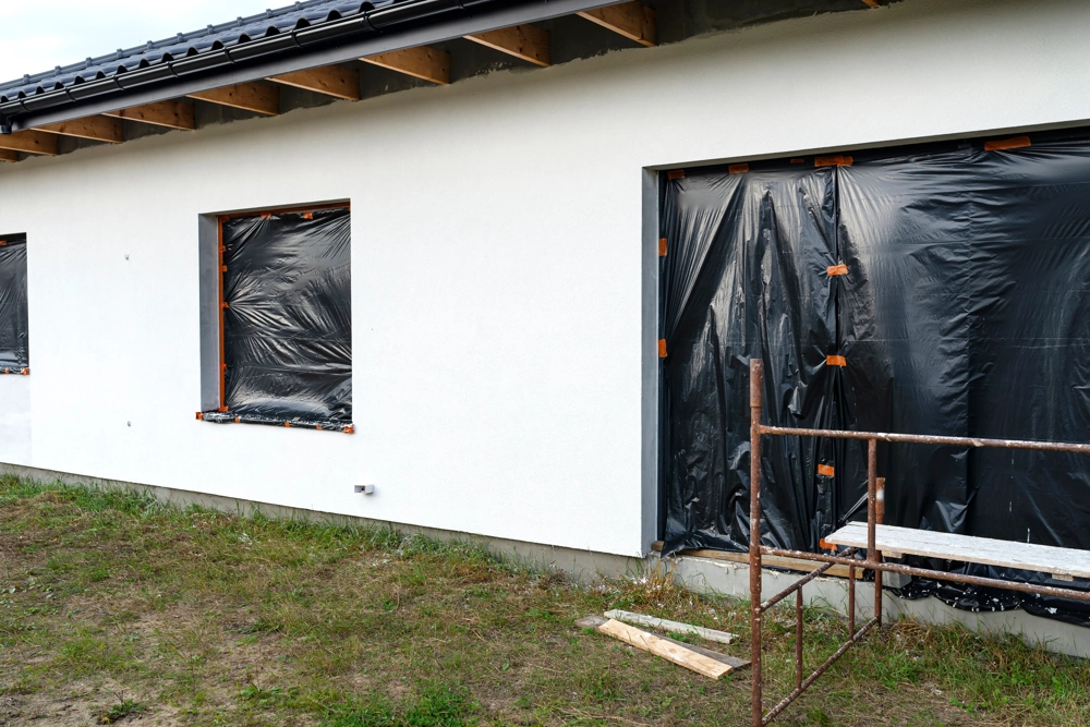 Elewacja domu w trakcie prac, biała ściana z rusztowaniem i oknami zabezpieczonymi czarną folią